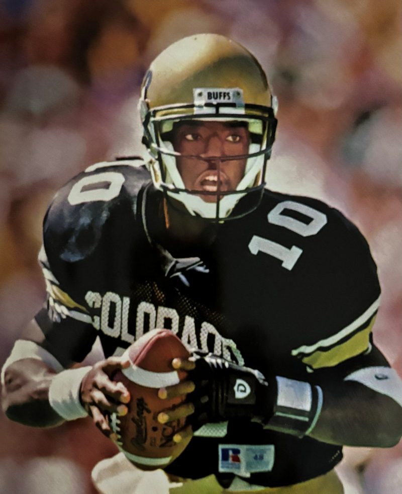 Football player in black and gold uniform holding a ball on the field.